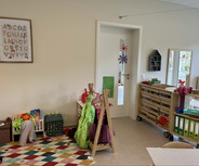 In einem hellen Raum sind Spielmaterialien und Kostüme für Kinder sichtbar. An der Wand hängt ein Alphabet-Plakat. Es gibt Regale mit Aufbewahrungskästen, einen Spiegel und eine Tür mit einem kleinen Fenster. Der Boden ist mit einem bunten, gemusterten Teppich bedeckt.