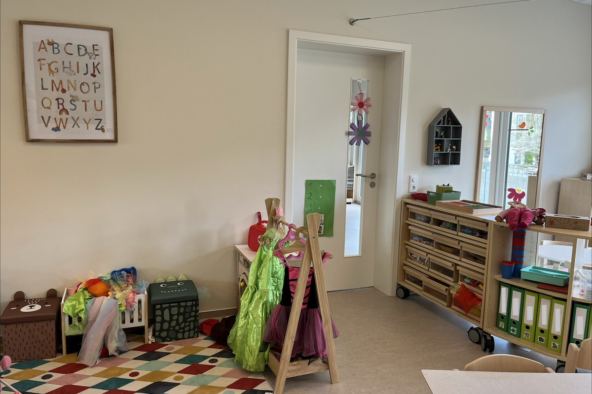 In einem hellen Raum sind Spielmaterialien und Kostüme für Kinder sichtbar. An der Wand hängt ein Alphabet-Plakat. Es gibt Regale mit Aufbewahrungskästen, einen Spiegel und eine Tür mit einem kleinen Fenster. Der Boden ist mit einem bunten, gemusterten Teppich bedeckt.