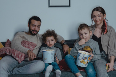 Eine Familie sitzt auf einer Couch und genießt einen Filmabend. Sie haben Popcorn in großen Schüsseln und schauen zusammen auf einen Fernseher. Die Eltern und zwei Kinder sind entspannt und lächeln. Kissen und eine neutrale Wand umgeben die Szene.