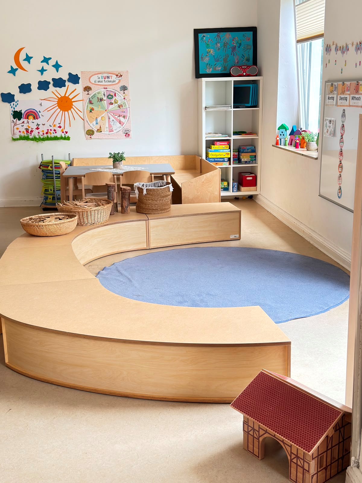 Ein heller Spielraum mit einer großen, geschwungenen Holzmöbelkombination, die kleine Tische und Sitzbereiche bietet. An der Wand hängen bunte Kinderzeichnungen. Ein blauer Teppich liegt auf dem Boden. Neben Fenstern sind Regale mit Spielzeug und Materialien.