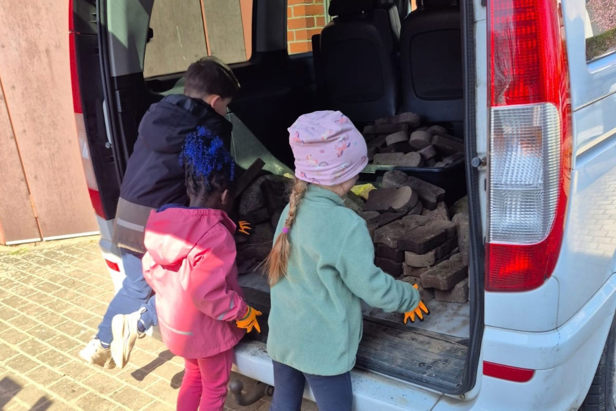 Drei Kinder stehen am offenen Kofferraum eines Lieferwagens und heben Steine oder Platten hinein. Sie tragen Handschuhe und scheinen beschäftigt zu sein.