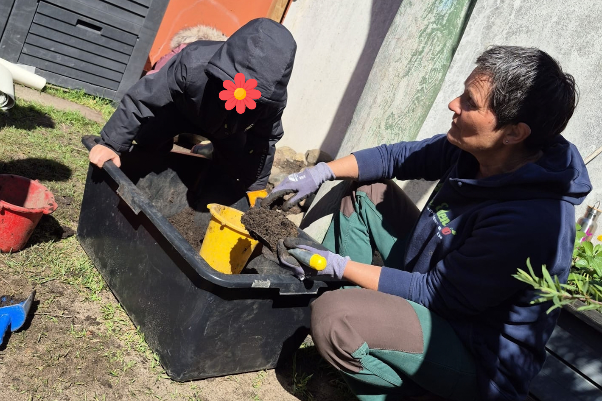 Eine Person mit grauen Handschuhen und blauer Jacke arbeitet mit einem Kind, das in einem schwarzen Behälter sitzt. Beide sind im Freien und beschäftigen sich mit Erde. Im Hintergrund sind verschiedene Gartenutensilien sichtbar.