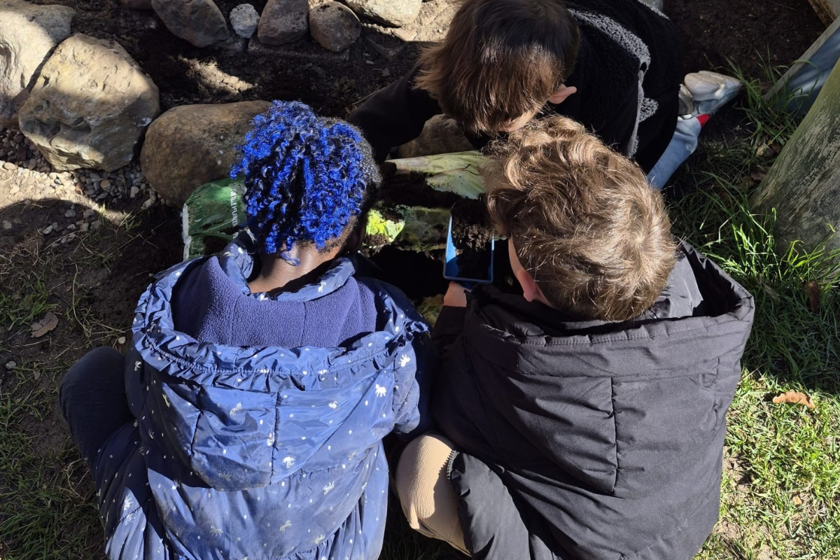 Drei Kinder, die auf dem Boden sitzen und an einer Pflanze oder einem Naturobjekt interessiert sind. Eines der Kinder hält ein Handy, um etwas zu fotografieren oder zu filmen. Sie sind umgeben von Steinen und Gras in einer natürlichen Umgebung.